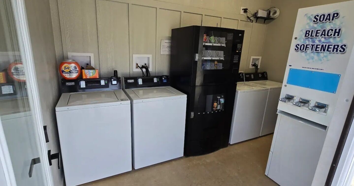 Ein Waschraum mit vier Waschmaschinen, einem schwarzen Kühlschrank sowie einer Spender für Seife, Bleichmittel und Weichspüler.
