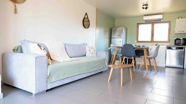 A modern living room with a sofa, a dining table with chairs, a kitchen area with a refrigerator and microwave, and a wall-mounted clock.