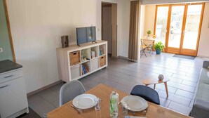 A dining area with a table set for two, a television on a white cabinet, and a sliding glass door leading to an outdoor area with a table and chairs.