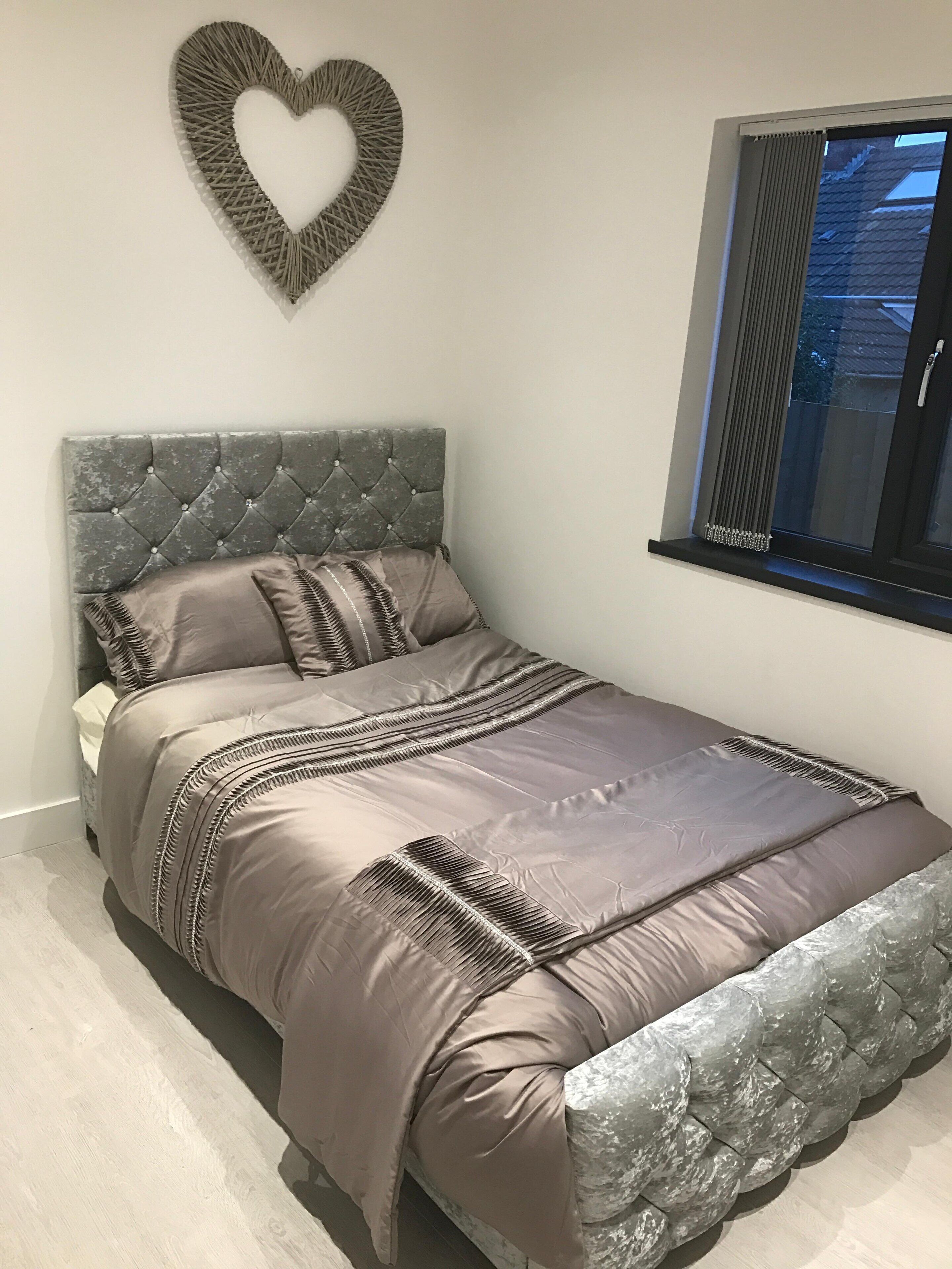 A bedroom with a tufted headboard, a heart-shaped wall decoration, and a window with blinds.