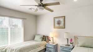 Une chambre à coucher comprenant un lit, deux tables de chevet, un ventilateur de plafond, une fenêtre avec des stores et un tableau encadré accroché au mur.
