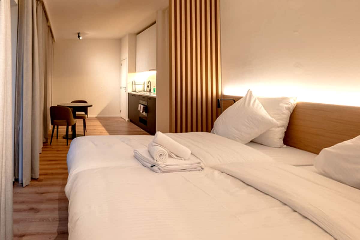 Una camera d'albergo con un letto rifatto, una scrivania con una sedia e una piccola cucina sullo sfondo.
