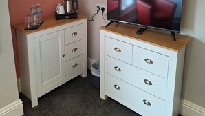 Una habitación pequeña con una televisión sobre un mueble de madera, un armario blanco con cajones, una cafetera y un cesto de basura.