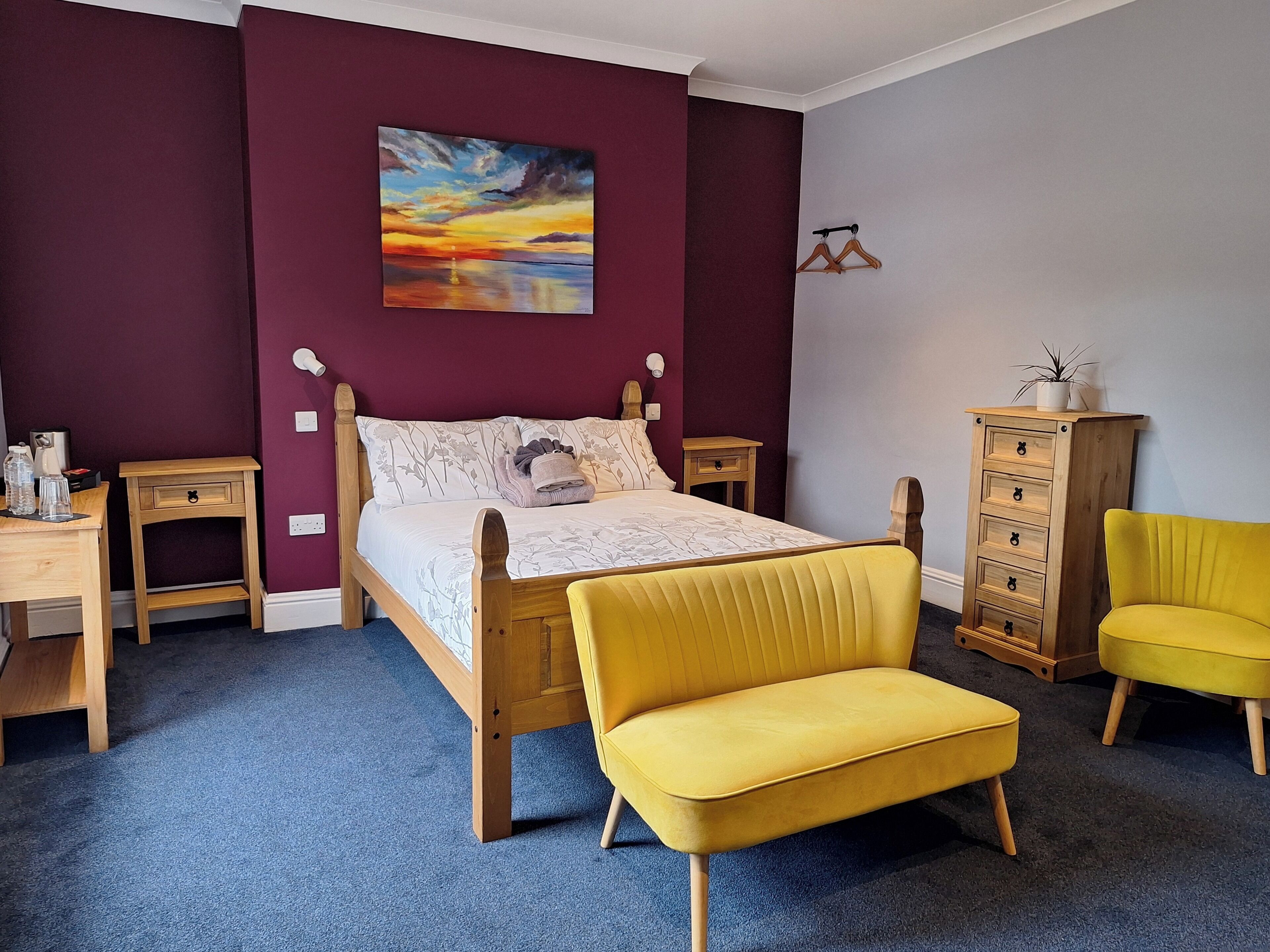 A bedroom with a bed, two nightstands, a wooden dresser, a yellow armchair, and a painting on the wall.