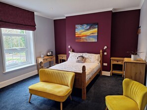 A bedroom with a bed, two nightstands, a wooden dresser, a yellow armchair, a window with a view, and a painting on the wall. - Room-comfortable and Spacious Double En- (Cranleigh)
