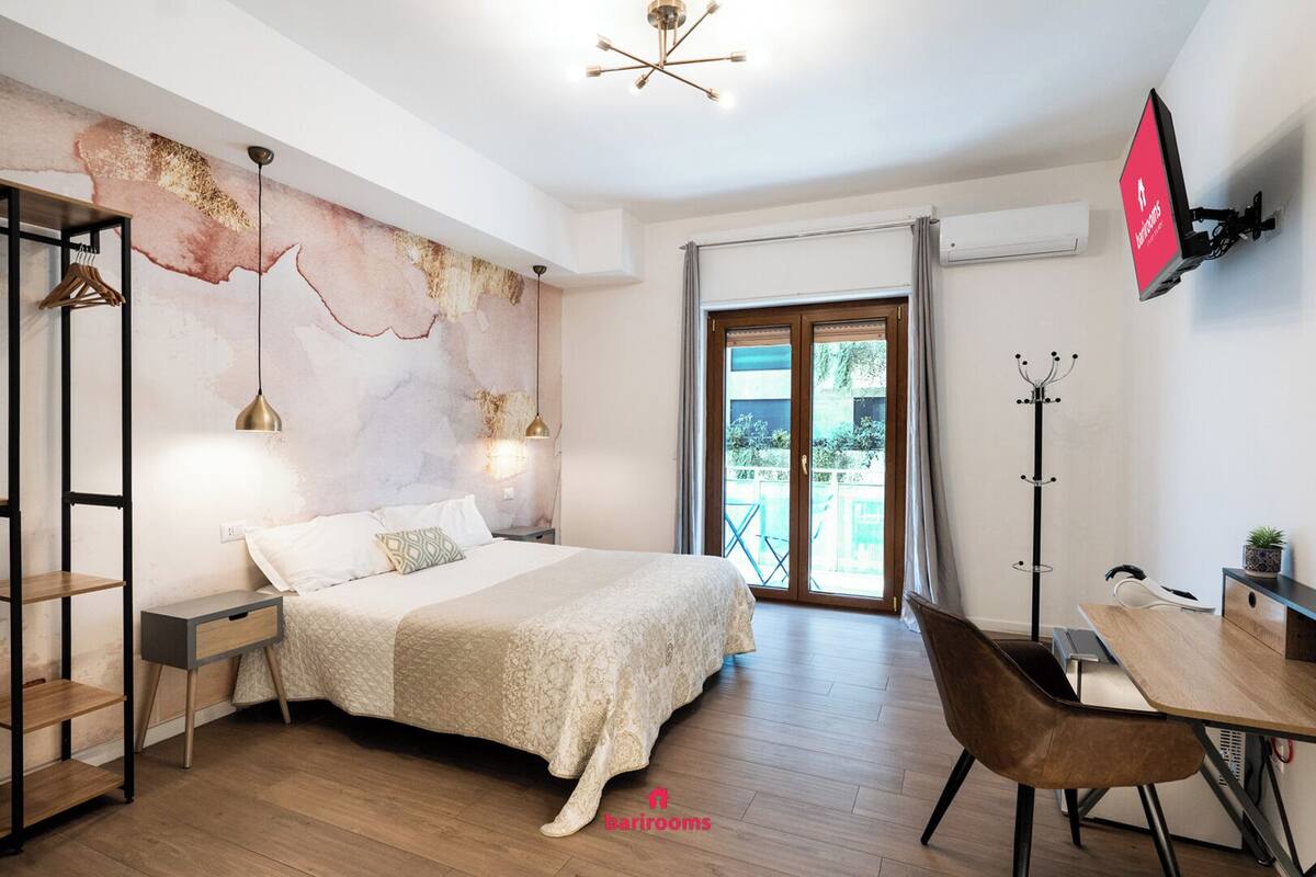 Un dormitorio moderno con cama, mesitas de noche, un escritorio y una silla. Hay un ventanal con cortinas y vistas a zonas verdes.