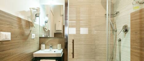 Une salle de bain moderne avec un meuble-lavabo en bois, un évier, deux distributeurs de savon, une serviette, un miroir, un sèche-cheveux et une douche en verre avec un pommeau de douche amovible.