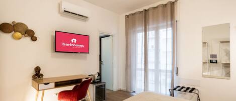 A hotel room with a bed, a desk with a chair, a television displaying 'barrirooms', an air conditioner, and a window with curtains.