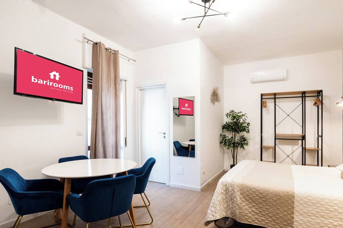 Una habitación de hotel moderna con una cama, una mesa redonda con sillas, un televisor mostrando 'barrirooms' y un cuadro abstracto grande en la pared.