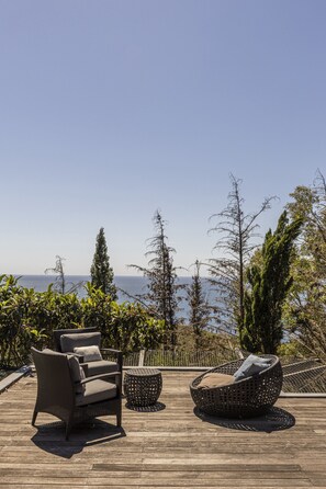 Un terrazzo in legno con mobili in vimini, tra cui una sedia e un tavolo rotondo, con vista panoramica su alberi e mare.
