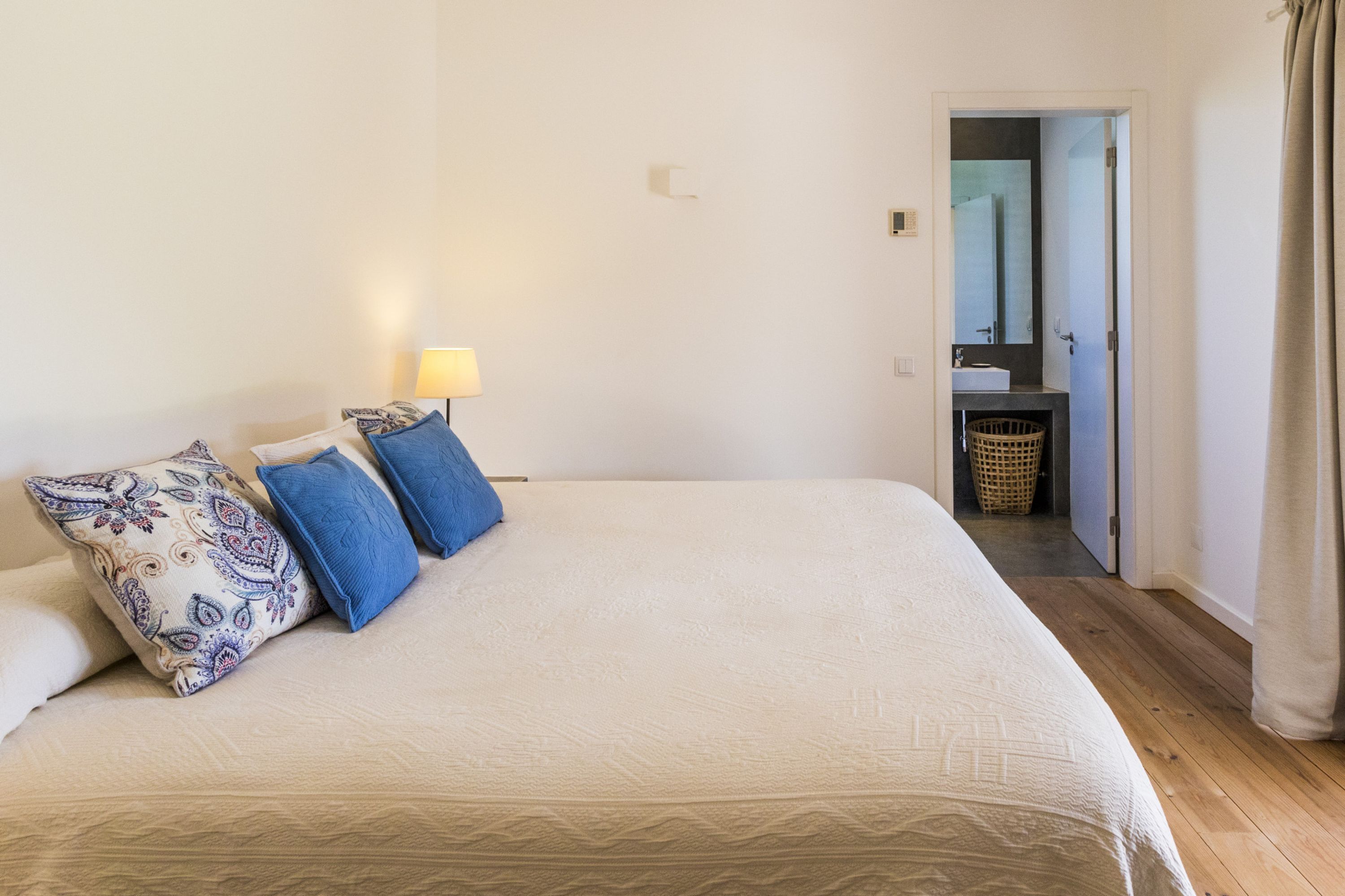 Een net opgemaakt bed met gestreepte kussens, een bedlampje en uitzicht op een badkamer met spiegel en wastafel.