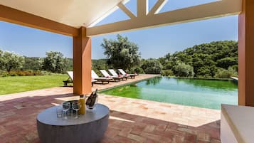 Relax by the serene pool surrounded by lush greenery and a clear sky, with comfortable lounge chairs and a shaded patio area.