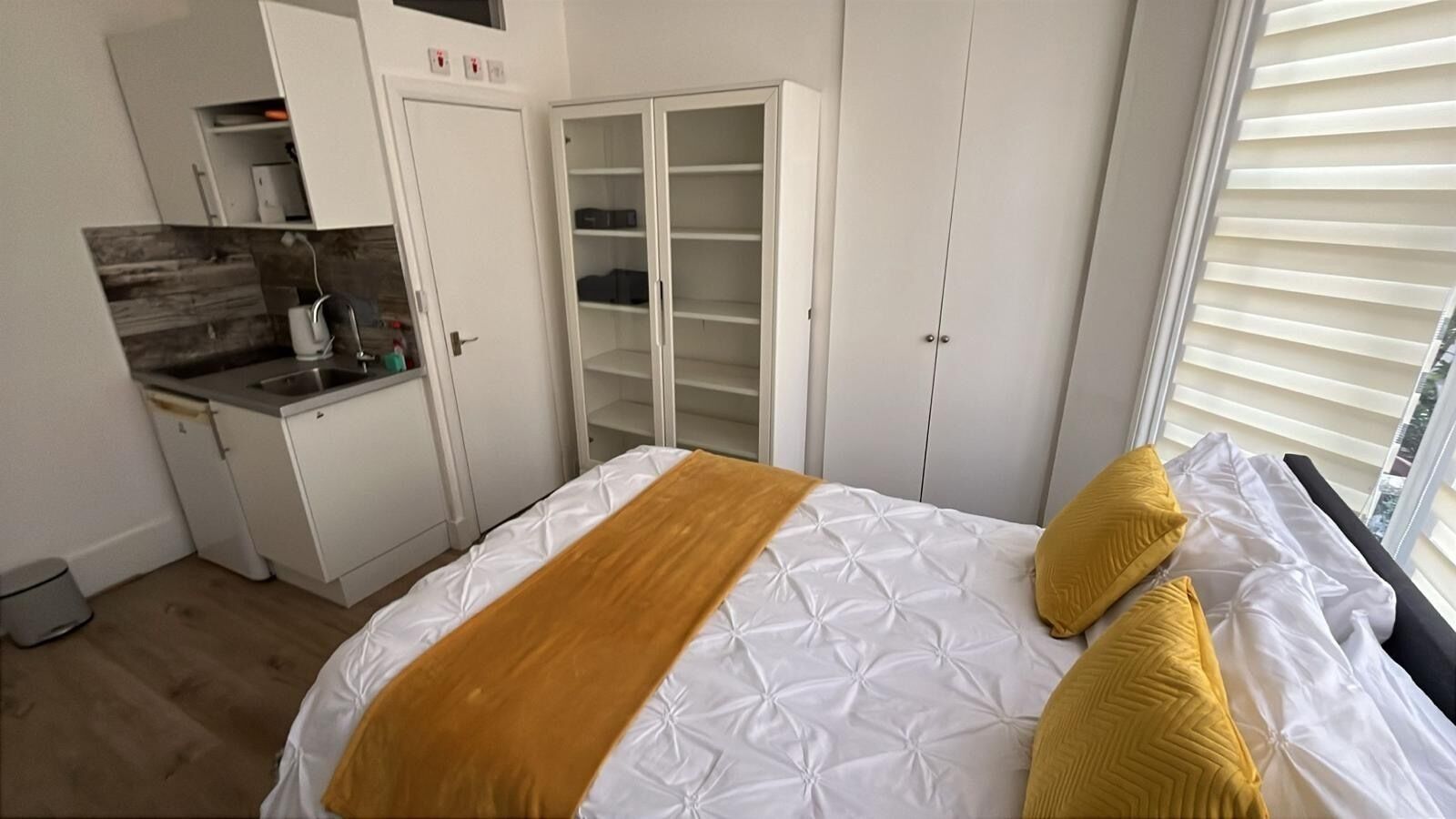 Ein Schlafzimmer mit Bett, Kleiderschrank, einer kleinen Küchenzeile und einem Fenster mit Jalousien.