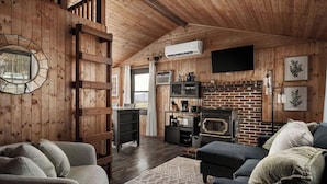 A cozy cabin interior with wooden walls, a brick fireplace, a flat-screen TV, and a comfortable seating area. - Cozy Mountain Cabin with Stunning Views, Modern Comforts & Fast WiFi (Bentonville)