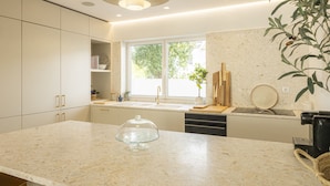 A modern kitchen with a large island, a window with a view, and a plant. - Contemporary Restelo Pool Haven by Cadenzalux (Lisbon)