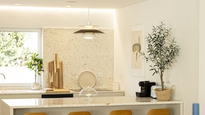 A modern kitchen with a central island, bar stools, a wooden dining table, and a view of the outdoors. - Contemporary Restelo Pool Haven by Cadenzalux (Lisbon)
