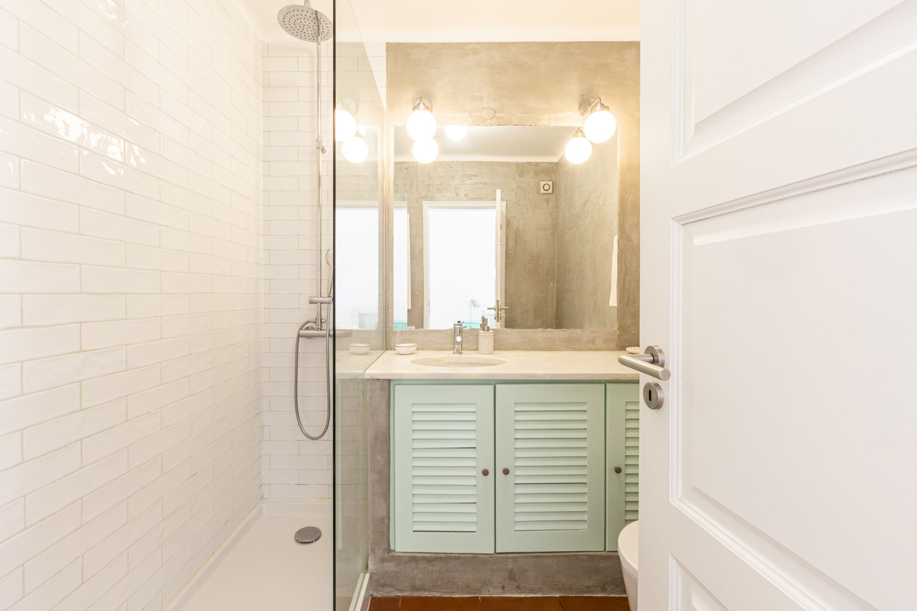 A bathroom with white tiled walls, a glass shower enclosure, a sink with a mirror, and light green cabinets.