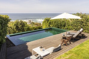 A private pool with a view of the ocean, wooden deck, lounge chairs, and a table with a glass of wine.