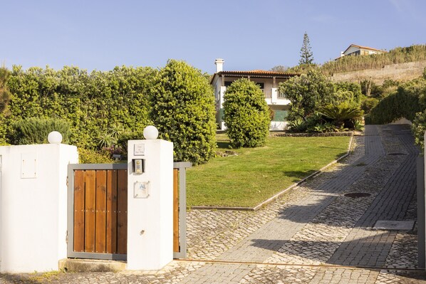 A gated entrance with a wooden gate, a paved pathway, and a house with a sloped roof and a garden in the background.