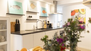 A modern kitchen with white cabinets, a black countertop, and various kitchen utensils. There are framed fish illustrations on the wall and a painting of fruits and vegetables.