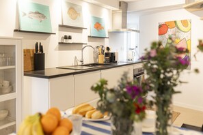 A modern kitchen with white cabinets, a black countertop, and various kitchen utensils. There are framed fish illustrations on the wall and a painting of fruits and vegetables. - Cascais Petit Bijou by Cadenzalux (Cascais)