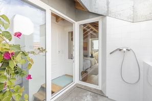 A modern bathroom with a glass door, a shower, and a visible bedroom with a bed and a nightstand.