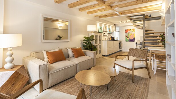 A modern living room with a sofa, coffee table, and chairs. There is a kitchen area with cabinets and a dining table. A staircase is visible in the background.