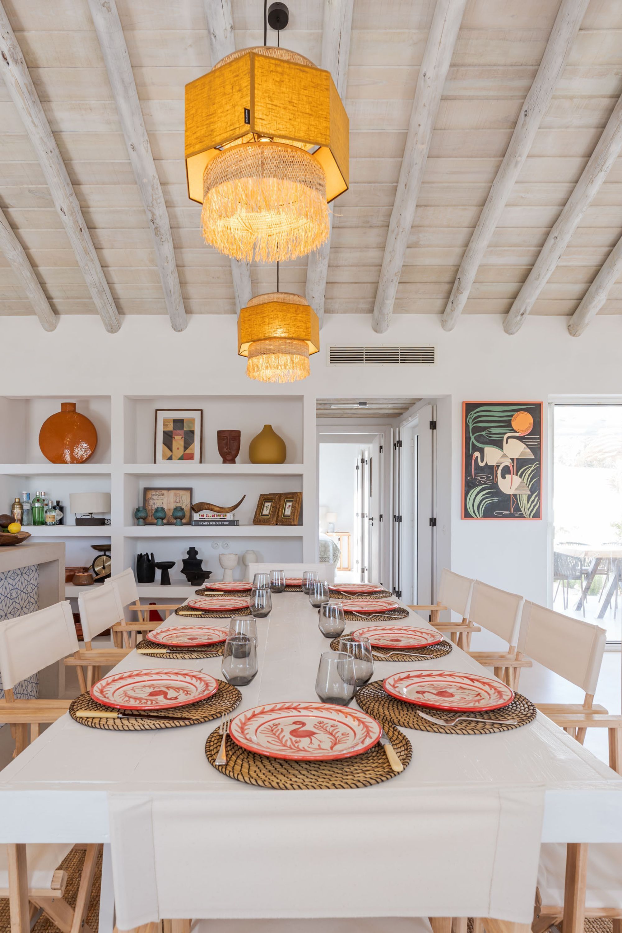 A dining area with a long table set for six, featuring woven placemats and red and white plates.
