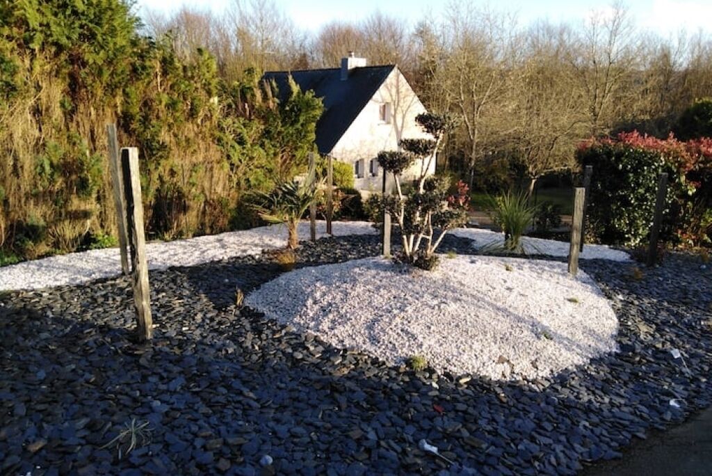 Une maison entourée de gravier et de petites plantes, avec des arbres en arrière-plan.