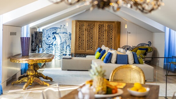 A modern loft bedroom with a high ceiling, a large window, a decorative wooden cabinet, a sofa with colorful cushions, and a dining table with fruit. - Chiado Rooftop Apartment by Cadenzalux (Lisbon)