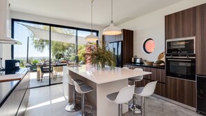 Een moderne keuken met een centraal eiland, barkrukken, een ingebouwde oven en een groot raam met uitzicht op een tuin.