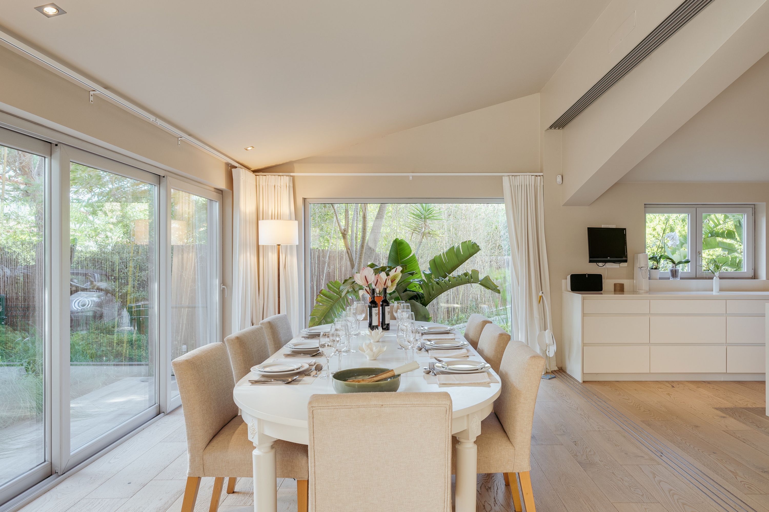 A modern dining room with a large table set for six, surrounded by large windows offering a view of greenery.