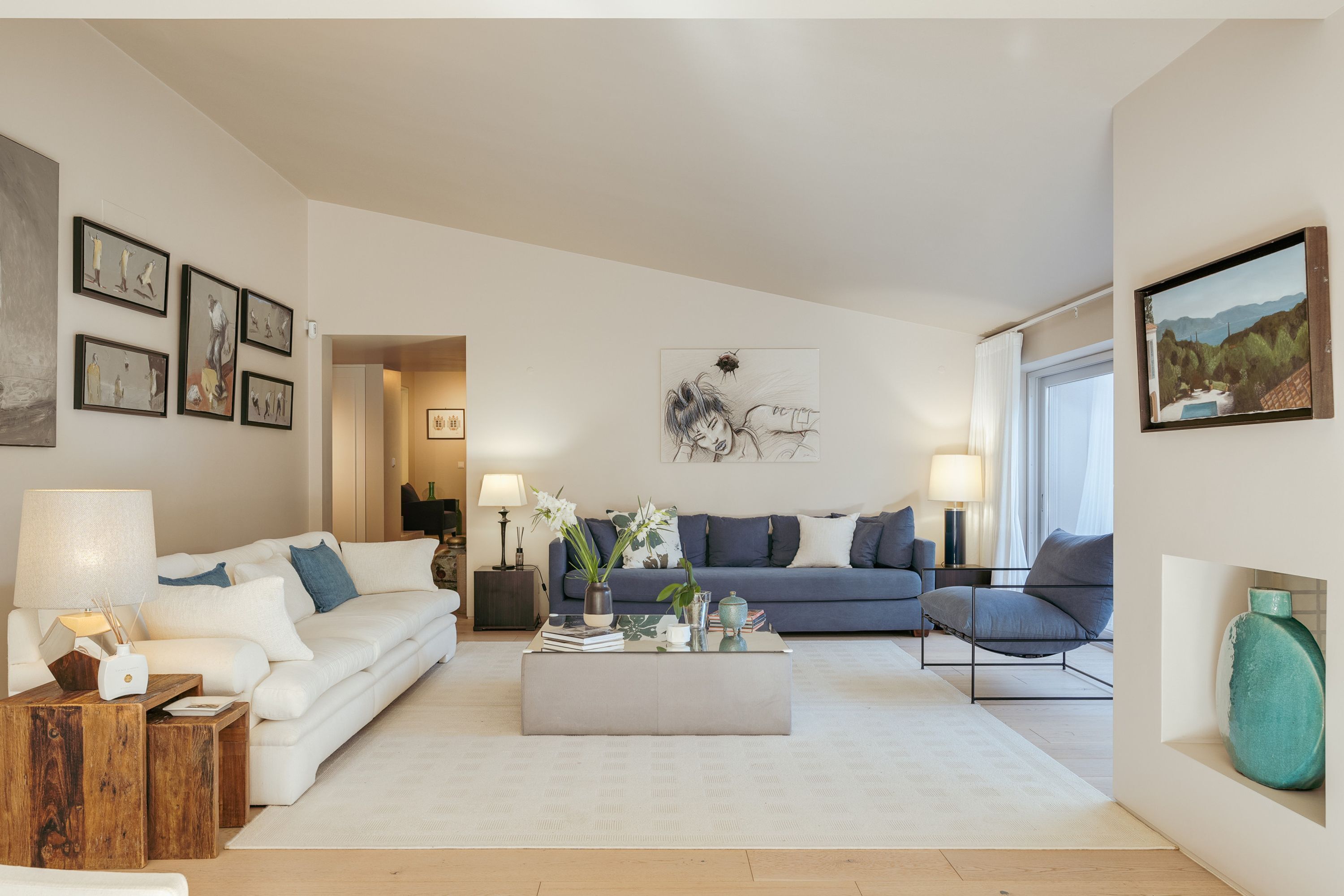 A modern living room with a white sofa, a blue sofa, and a grey coffee table. There are framed pictures on the walls and a painting above the blue sofa.
