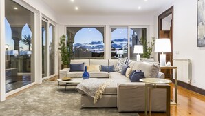 Un salon moderne avec un grand canapé d’angle, une table basse et une table d’appoint. On y trouve deux lampes, une plante en pot et une vue sur l’extérieur grâce à de grandes fenêtres.