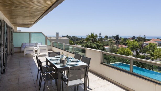 Un balcón con una mesa de comedor dispuesta para cuatro personas, con vistas a una piscina y una panorámica de la playa y edificios circundantes.