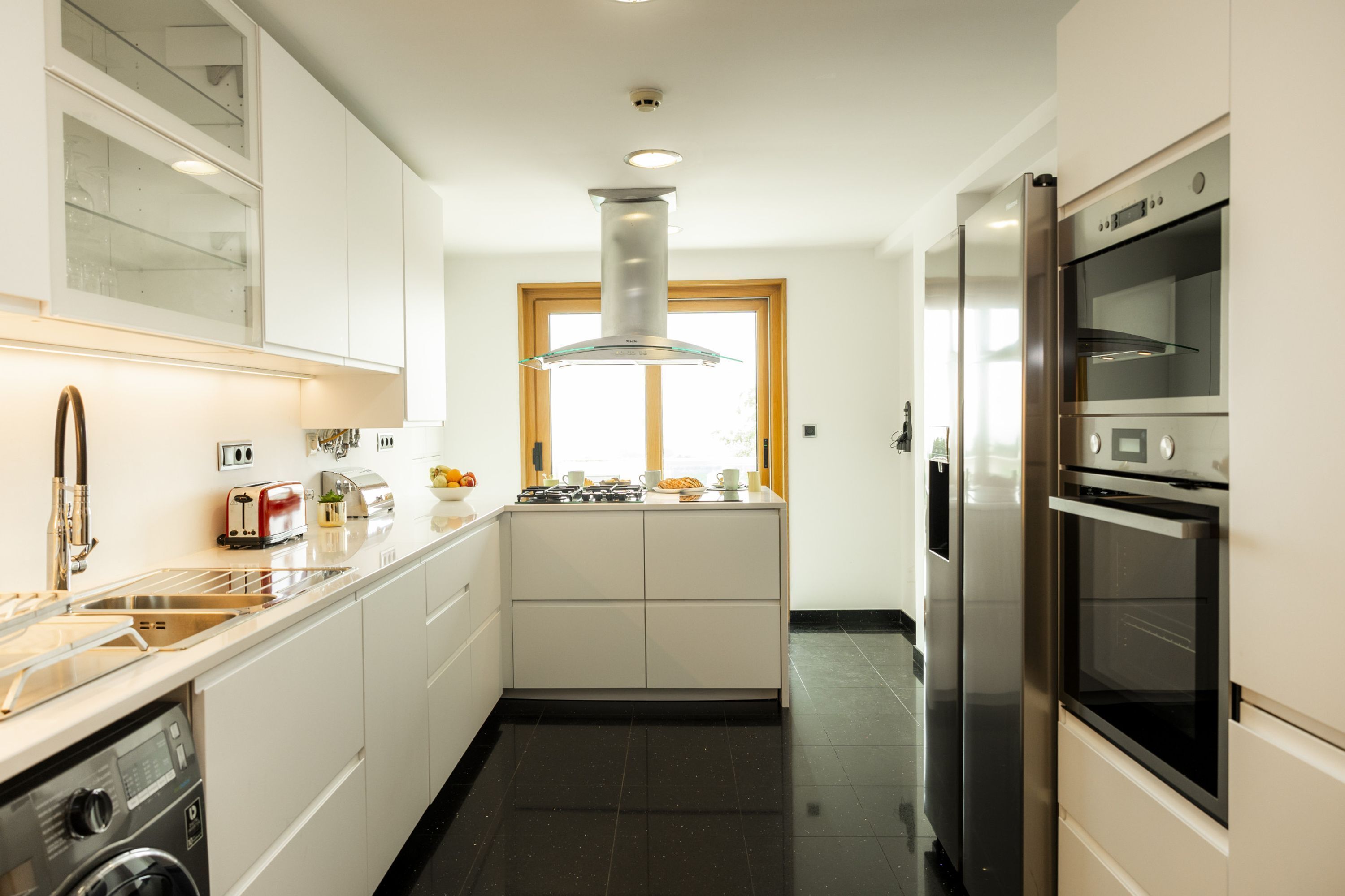 A modern kitchen with white cabinetry, stainless steel appliances, a built-in oven, and a window providing natural light.
