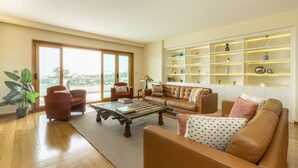 Un salón amplio con un gran ventanal, un sofá de cuero, una mesa de centro y una estantería con objetos decorativos.