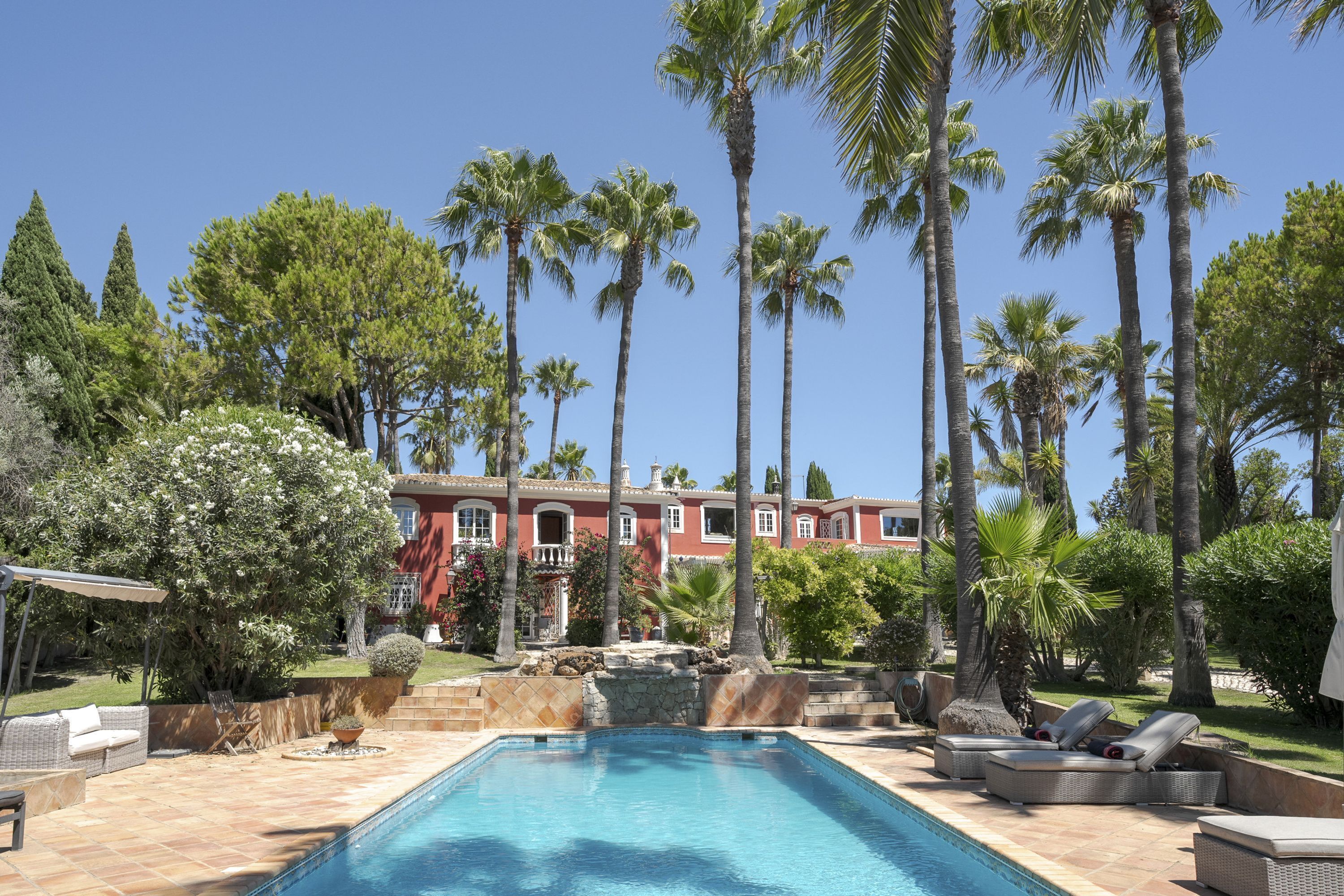 A swimming pool with lounge chairs, surrounded by palm trees and a red building in the background.