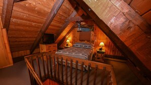 Een gezellig slaapkamer op zolder met een schuine houten plafond, een bed met beddengoed met een patroon, twee bedlampen en een houten stoel.