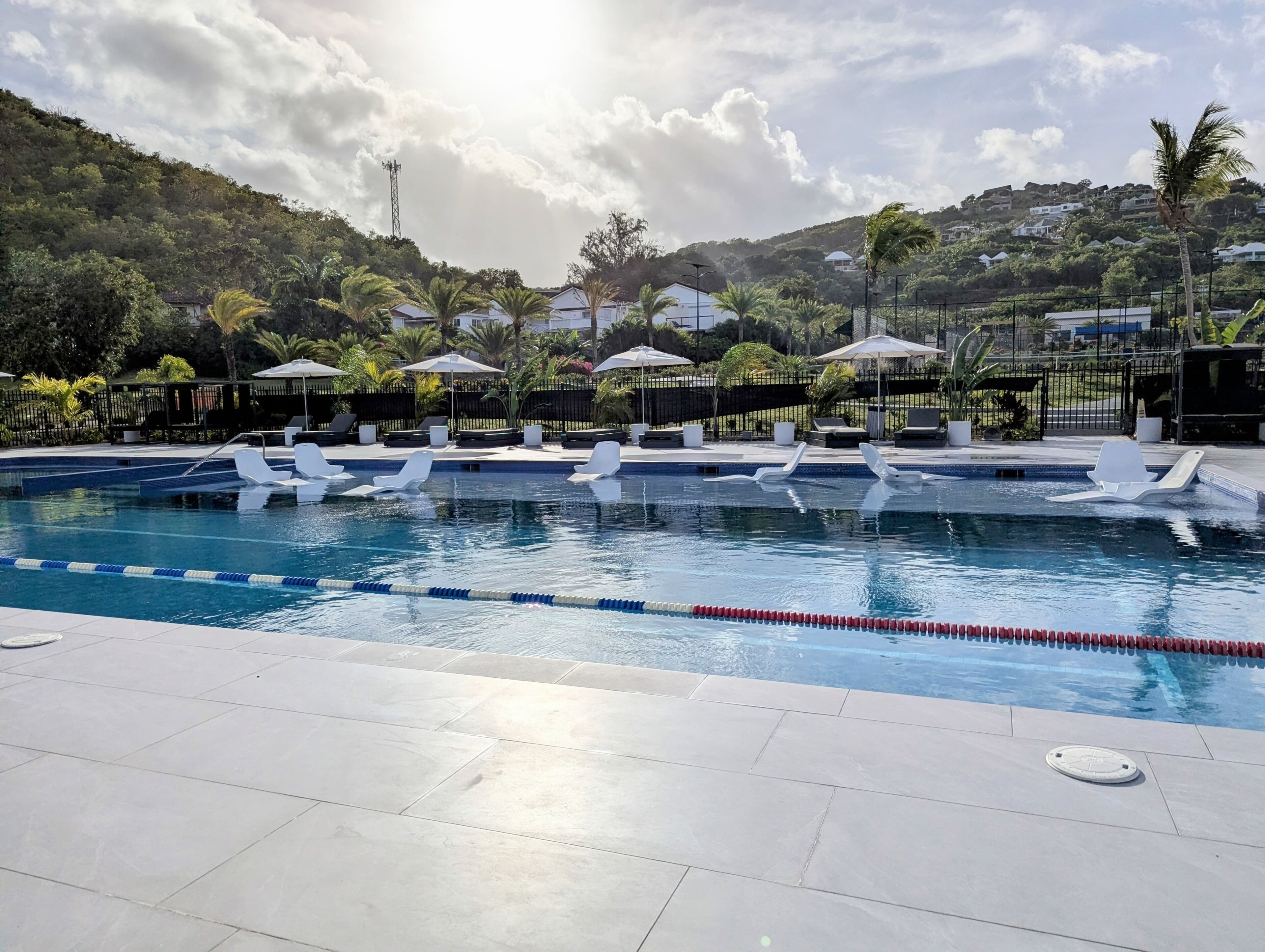 Una piscina con sillas de descanso y sombrillas, rodeada de vegetación y casas.