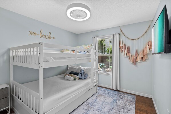 A bunk bed with a white frame and a ceiling fan, a wall-mounted TV, and a window with blinds.