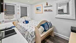 A bedroom with a bed, a desk, a chair, a window, and a shelf with decorative items.