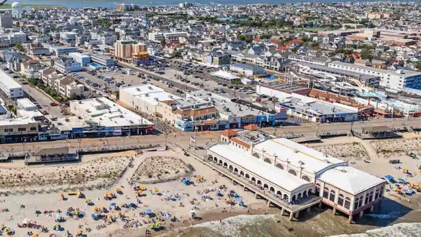 An aerial view of a coastal city with a large beach area, numerous buildings, and a prominent white structure with arches.