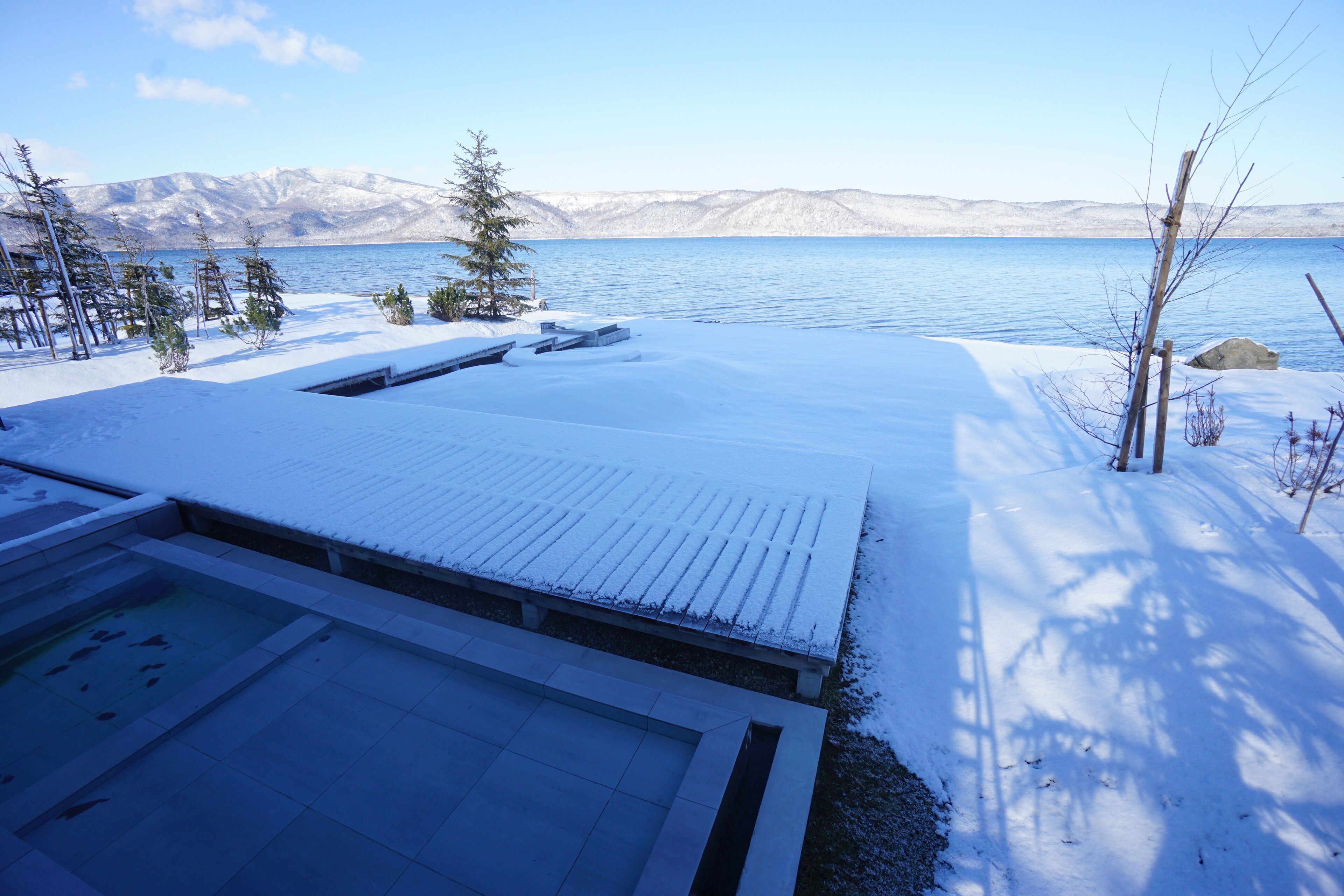 Un espace extérieur enneigé comprenant une piscine, une terrasse en bois et une vue sur un lac et des montagnes.