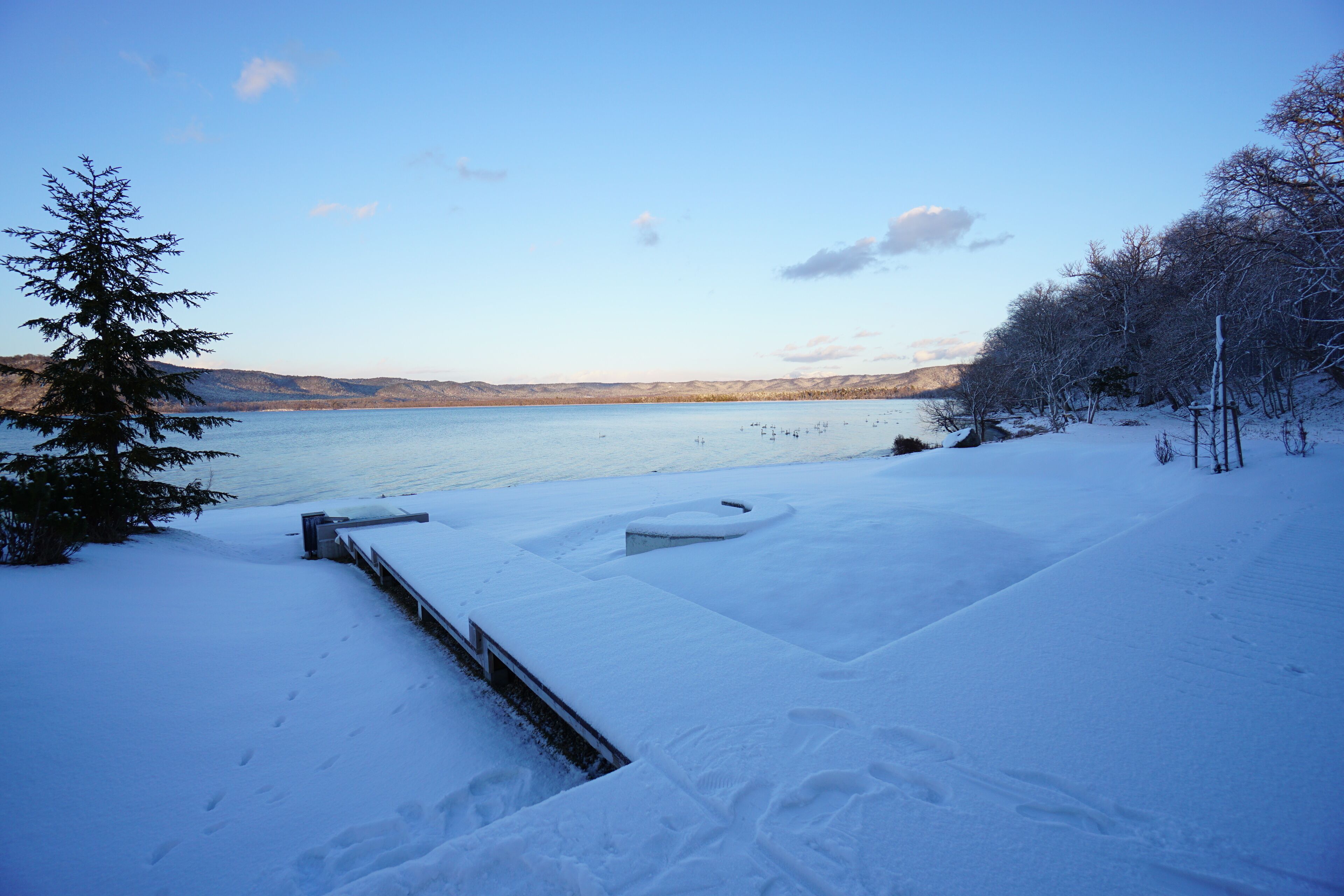 Un paysage enneigé avec un lac, une terrasse en bois et un sentier recouvert de neige menant à un banc.