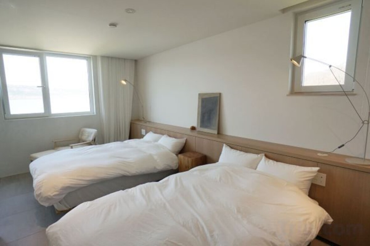Una habitación de hotel con dos camas, una mesita de noche, una silla, un ventanal y una lámpara.