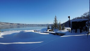 A snow-covered landscape with a lake, a wooden deck, and a building with a garage. - Sansui Lakeside Villa (Teshikaga)