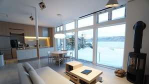 A modern living room with a sofa, a coffee table, and a fireplace. There is a kitchen area with a dining table and chairs. Large windows provide a view of a snowy landscape. - Sansui Lakeside Villa (Teshikaga)