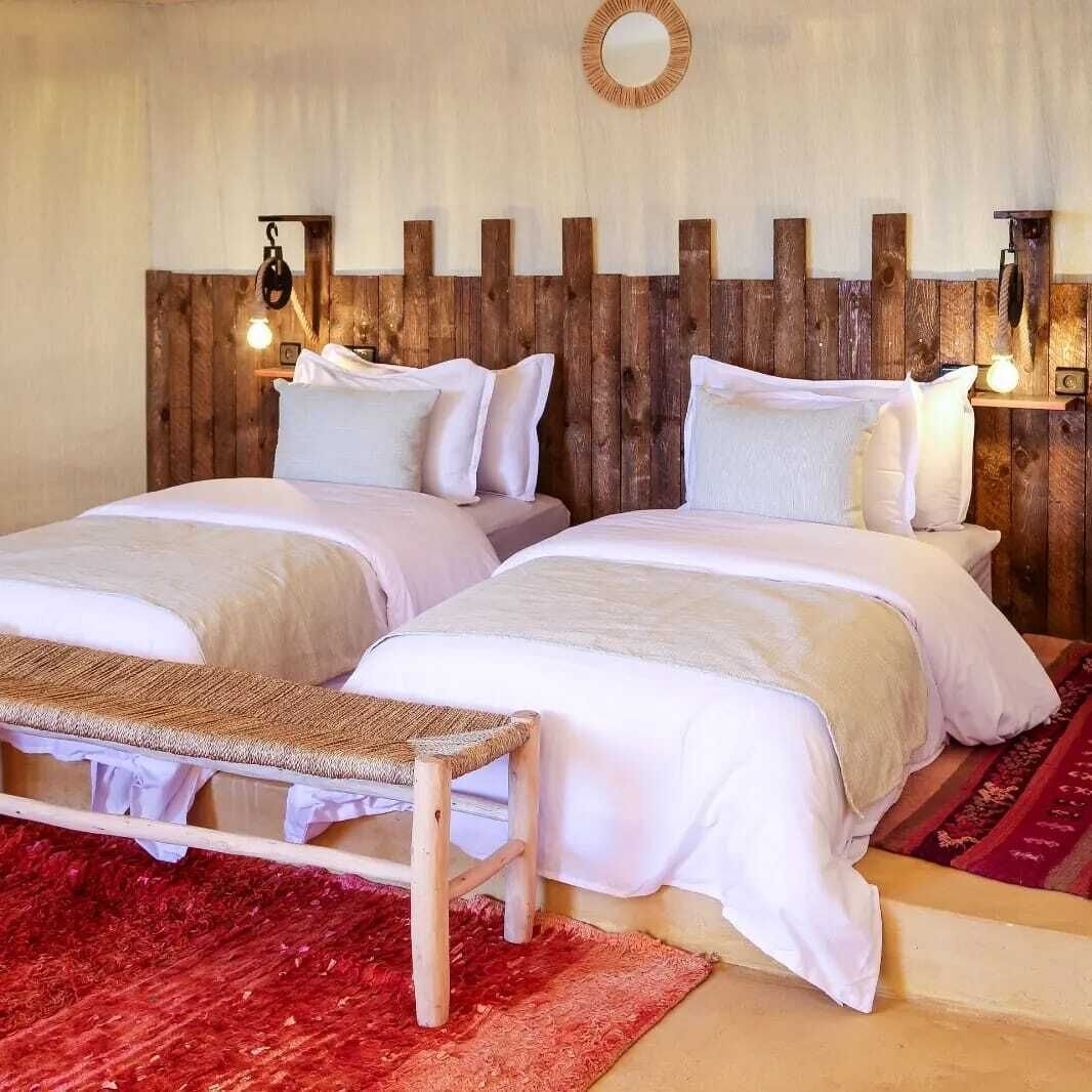 A double room with two beds, a bench, a red rug, and a wooden headboard.