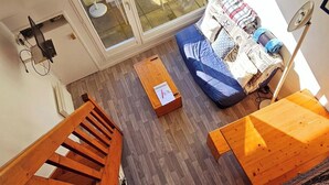 A room with a wooden floor, a bed with a blue blanket, a small wooden table, and a window with a view of the outside. - Bright apartment between port and beaches (Arzon)